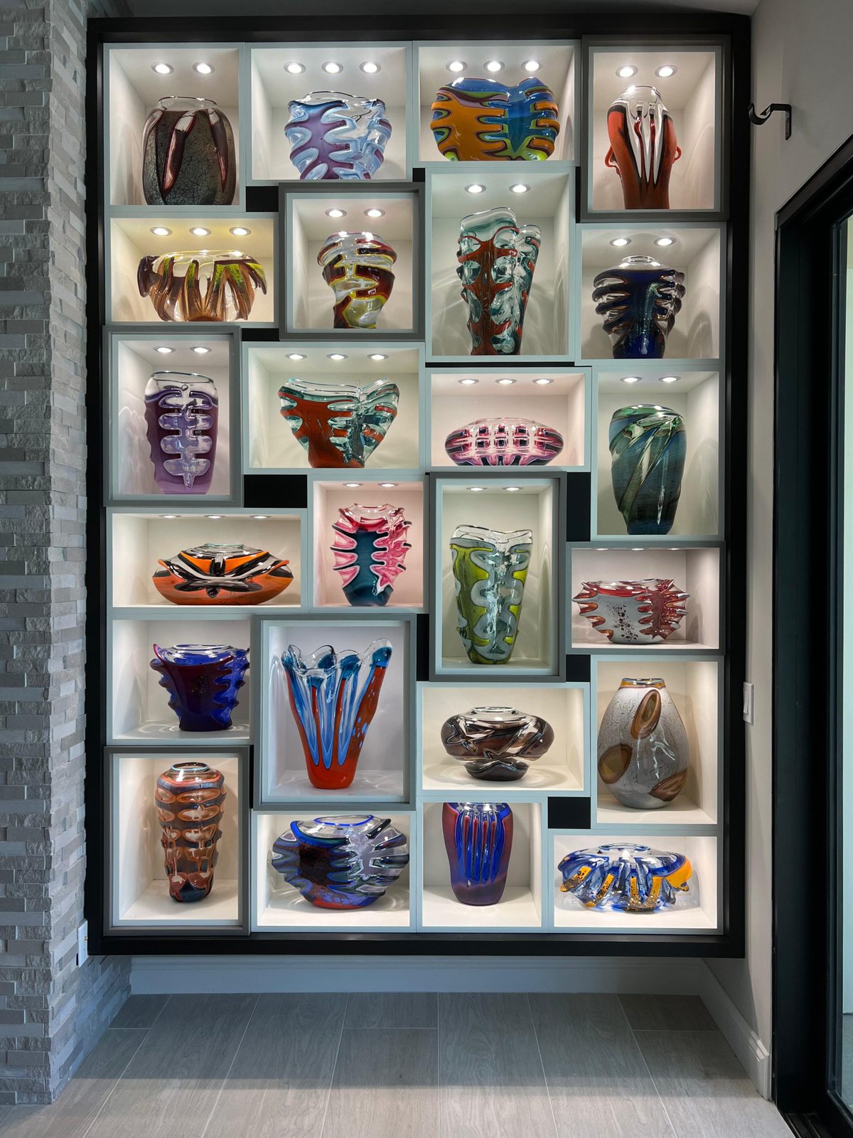 Floor-to-ceiling wall of lit display niches holding colorful glass vases