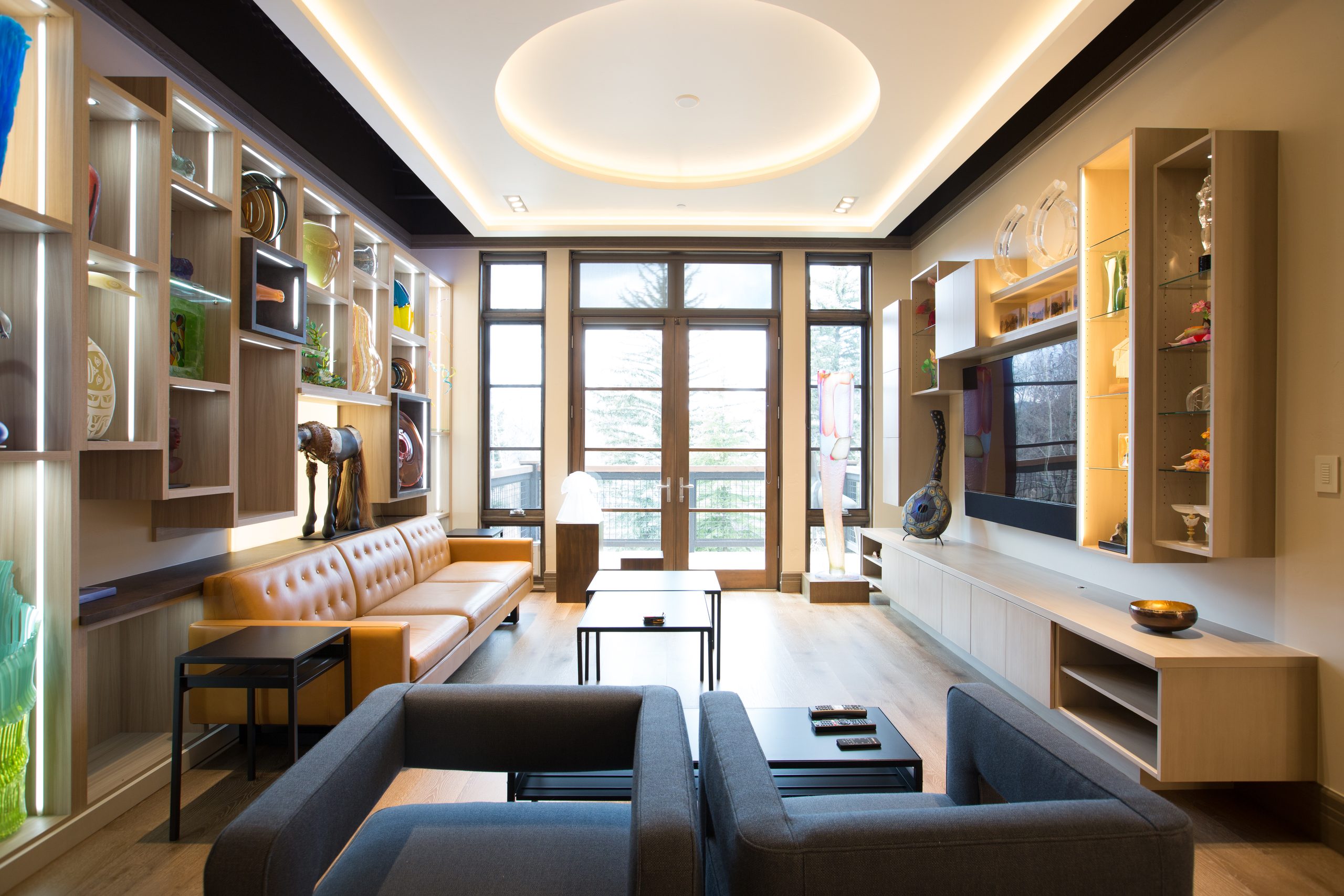 Living room with lit custom display shelving, a tan leather sofa and two grey armchairs